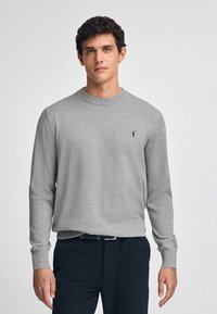 Maglione grigio a maniche lunghe con scollo rotondo e polsini a coste. Presenta un piccolo logo nero sul lato sinistro del petto. Indossato con pantaloni scuri.