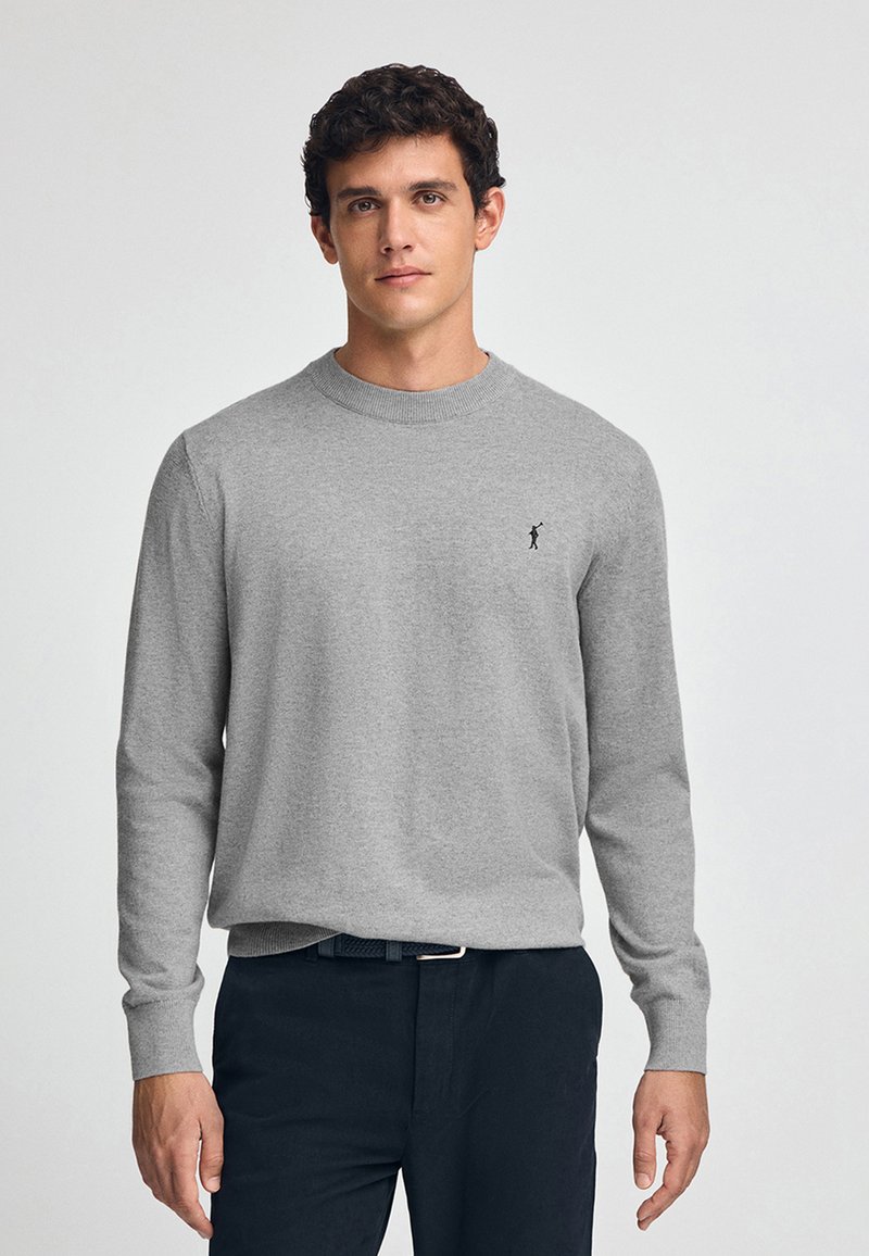 Maglione grigio a maniche lunghe con scollo rotondo e polsini a coste. Presenta un piccolo logo nero sul lato sinistro del petto. Indossato con pantaloni scuri.