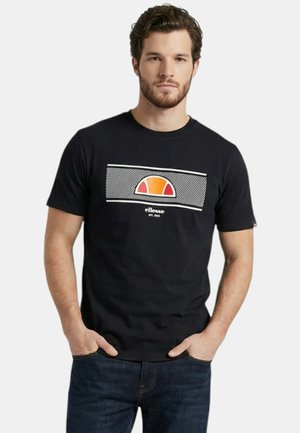 Homme portant un t-shirt noir de la marque ellesse avec un logo en demi-cercle orange et rouge, debout les mains dans les poches de son jean sur fond blanc.