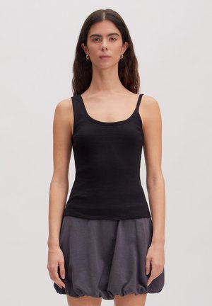 Vrouw met lang donker haar die gouden oorringen draagt, een zwart topje en een donkergrijze ballonrok, staande voor een effen achtergrond.