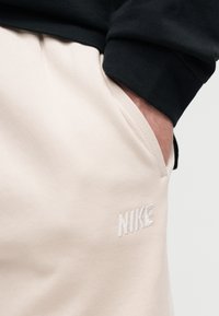 Χέρι στην τσέπη μπεζ φόρμας Nike με κεντημένο λογότυπο, συνδυασμένη με μαύρο μακρυμάνικο τοπ.