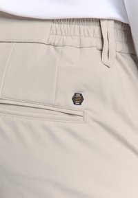 Beige broek van lichtgewicht stof met een elastische tailleband, voorzien van een zijzak en een hexagonaal zwart logo accent.