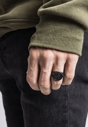 Fekete gyűrű, mintázott felülettel, kézen viselve, olíva zöld pulóver és fekete farmernadrág kíséretében. Az anyag texturált hatású.