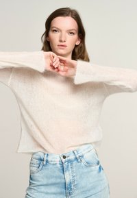 Maglione leggero e trasparente di colore bianco con una vestibilità oversized e maniche ampie, abbinato a pantaloni corti in denim azzurro. Materiale in maglia morbido e dalla consistenza particolare.