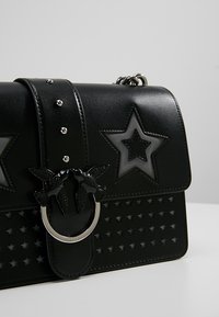 Borsa nera con forma rettangolare, caratterizzata da fori a forma di stella, hardware in metallo a forma di uccello e una texture in pelle liscia. Inclusi dettagli in argento.