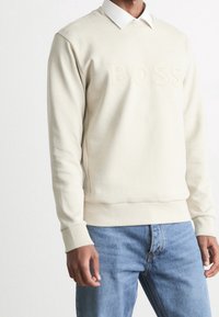 Beige sweatshirt med präglad "BOSS" logotyp, rund hals, och ribbade ärmslut, lager på lager över en vit skjorta med krage, kombinerad med jeans i denim.