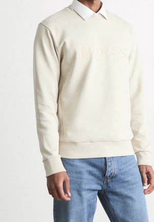 Felpa beige con logo "BOSS" in rilievo, collo rotondo e polsini a coste, indossata sopra una camicia bianca con colletto, abbinata a jeans in denim.