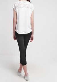 Top blanco con detalle de encaje en la espalda, combinado con leggins negros y zapatillas plateadas sin cordones. La parte superior tiene mangas cortas y un dobladillo curvado.