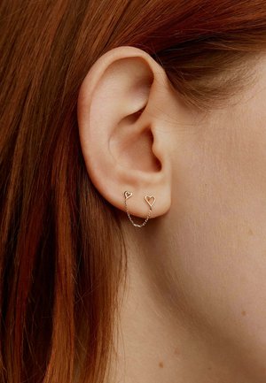 Pendientes en tono dorado con tachuelas en forma de corazón conectadas por una delicada cadena, llevados en la oreja de un modelo con un fondo de cabello rojo.