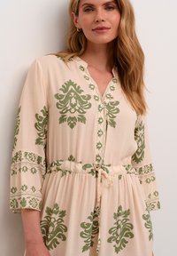 Long-sleeve beige dress featuring green floral patterns, a buttoned neckline, and an adjustable drawstring waist for a relaxed fit.