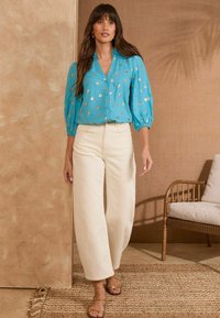 Una blusa turchese con bottoni e pois dorati, maniche a sbuffo, abbinata a pantaloni beige a vita alta e pantaloni larghi, con sandali dorati con strappi.