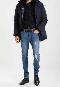 Giacca puffa blu navy con cappuccio foderato in pelliccia sopra una maglietta nera con piccoli motivi blu. Indossata con jeans blu strappati e stivali eleganti neri.