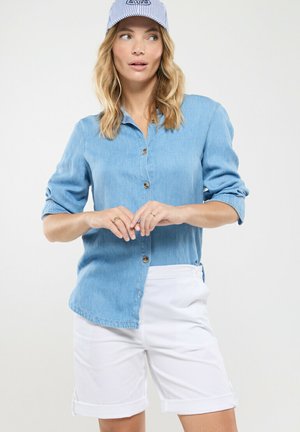 Femme portant une chemise bleu clair boutonnée avec les manches retroussées, un short blanc retroussé et une casquette rayée bleue et blanche, regardant sur le côté.