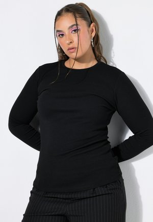 Frau mit langen Haaren und zwei dünnen Zöpfen, trägt rosa Augen-Make-up, schwarzes Langarmshirt und schwarze Nadelstreifenhose, steht mit den Händen hinter dem Rücken.
