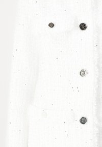 White tweed jacket featuring metallic buttons, a textured fabric with subtle shimmer, and fringe detailing along the edges.