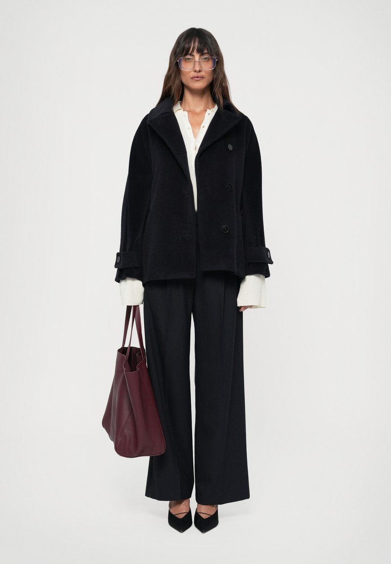 Casaco de lã preto com colarinho em bico, frente de botão duplo e mangas largas. Combinado com uma blusa branca, calças risca de giz e uma mala borgonha.