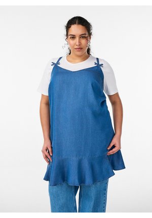 Femme portant une robe sans manches en denim bleu par-dessus un t-shirt blanc et un jean bleu, debout devant un fond blanc uni.