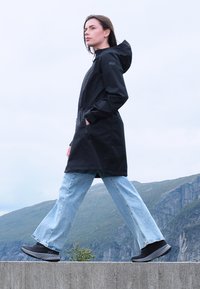 Mujer caminando sobre un borde de concreto, vistiendo un impermeable negro con capucha, jeans azul claro y zapatillas negras, con montañas al fondo.