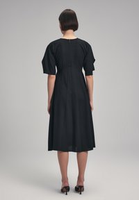 Musta midi-mekko lyhyillä, rakenteellisilla hihoilla, istuvalla bodysuitilla ja levenevällä hameella, jossa on takana vetoketjulukko. Silkkinen kankaan pinta.