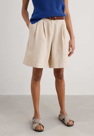 Pantaloni corti in lino beige con vestibilità rilassata, dotati di pieghe e tasche laterali. Abbinati a un top aderente blu e sandali grigi.