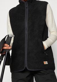 Schwarze Fleece-Weste mit hohem Kragen, voller Frontreißverschluss und zwei Seitentaschen. Verfügt über ein kleines Markenlogo auf der linken unteren Seite.