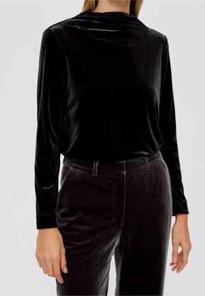 Žena oblečená v černém sametovém topu s dlouhým rukávem a odpovídajících černých sametových kalhotách, stojící před jednobarevným světlým pozadím.