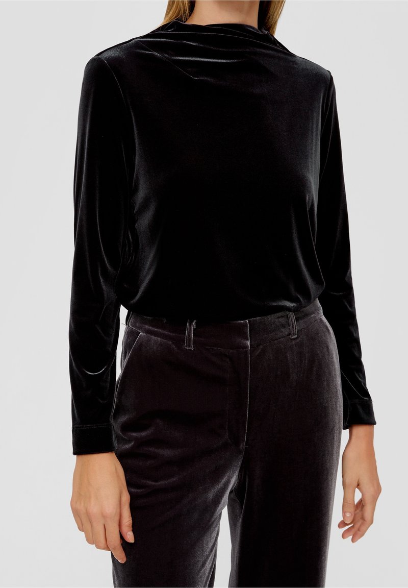 Femme portant un haut en velours noir à manches longues et un pantalon en velours noir assorti, debout devant un fond clair uni.