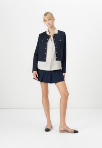 Vrouw staat rechtop en draagt een marineblauwe cropped jas, een beige blouse, een marineblauwe geplooide minirok en beige slingback flats met zwarte teentips.