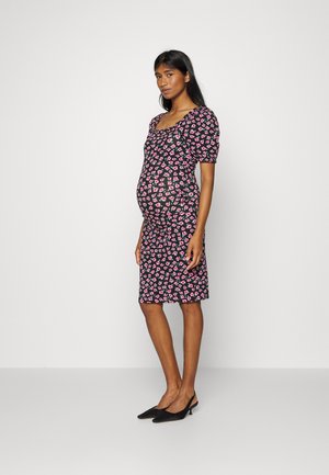 Vestido de maternidade com mangas curtas, tecido preto coberto por pequenos padrões florais em rosa, silhueta ajustada, até ao joelho, combinado com sapatos de salto pretos.