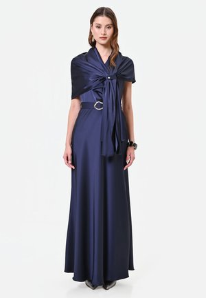 Femme portant une longue robe en satin bleu marine avec un châle assorti noué à la poitrine, une ceinture avec une boucle argentée et des talons noirs.