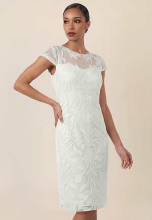 Frau trägt ein weißes knielanges Kleid mit durchsichtigen kurzen Ärmeln und großer strukturierter Blumenstickerei auf beigem Hintergrund.