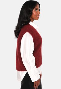 Burgunderfarget strikket vest laget av mykt materiale, har en ermeløs design, bæres over en hvit skjorte med knapper, og har en avslappet passform.