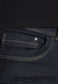 Jeans in denim blu scuro con una tasca frontale più piccola, caratterizzati da cuciture dorate a contrasto e dettagli hardware in metallo. Top nero a coste sopra.