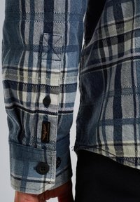 Manica di camicia a quadri blu e bianchi con polsini a bottone, caratterizzata da trame contrastanti e un'etichetta con logo vicino al polsino per l'identificazione.