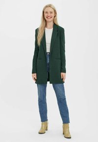 Grüner Blazer mit maßgeschneidertem Design, zwei vorderen Taschen, getragen über einem weißen Hemd, kombiniert mit blauen Jeans und strukturierten beigen Absätzen.