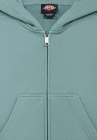 Φόρμα με φερμουάρ σε ανοιχτό πράσινο χρώμα, με τσέπη καγκουρό, φτιαγμένη από απαλό βαμβακερό ύφασμα· διαθέτει ασημένιο φερμουάρ και ετικέτα λογότυπου στο εσωτερικό του γιακά.