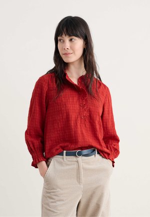 Chemise rouge légère à boutons avec un col volé et un plastron, manches longues avec des poignets ajustés ; assortie à un pantalon en velours côtelé beige et une ceinture bleue.