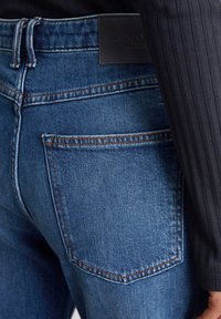 Jeans in denim blu dettagliati con cuciture arancioni, dotati di una tasca posteriore e di un'etichetta in pelle nera del marchio in vita.