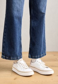 Baskets blanches avec des accents beige, des dessus texturés et un détail en corde sur la semelle, portées avec un pantalon en jean bleu à jambes larges.