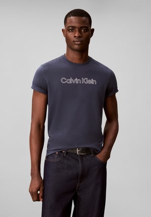 Jeune homme portant un t-shirt Calvin Klein bleu foncé et un jean sombre, debout avec une main dans la poche sur un fond gris uni.