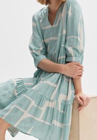 Femme assise, portant une robe vert menthe avec un motif géométrique blanc et des manches trois-quarts, reposant un bras sur une surface en bois.