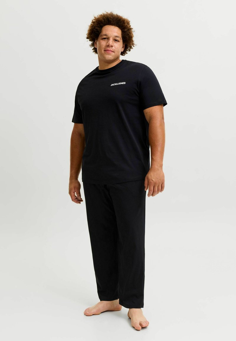 Homme debout pieds nus portant un t-shirt noir JACK & JONES et un pantalon noir sur un fond clair uni.