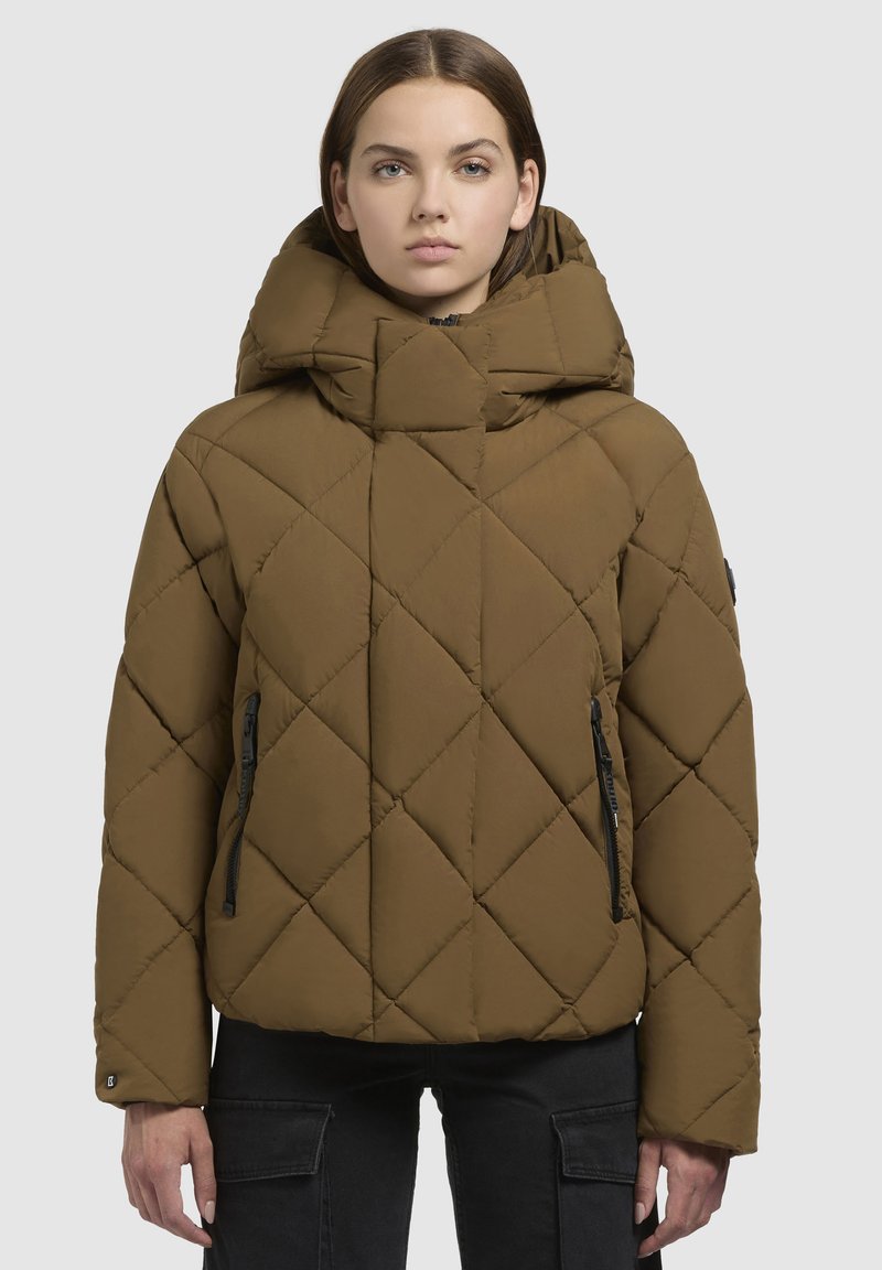 Giacca a vento imbottita marrone, con un ampio cappuccio, motivo di cuciture diagonali e tasche con zip. Il materiale sembra morbido e imbottito.