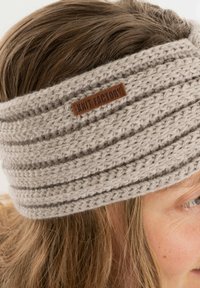 Lichtgrijze gebreide haarband van gestructureerd garen met horizontale ribbels en een bruine leren label met de tekst "KNIT FACTORY."