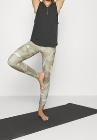 Czarny prążkowany top na ramiączkach oraz beżowe legginsy w kamuflaż. Legginsy mają gładką teksturę i półprzezroczyste wykończenie, zestawione z czarną matą do jogi.