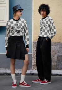 Chemises à manches longues à motif damier noir et blanc avec impression graphique, associées à une jupe plissée noire et un pantalon noir ample. Baskets rouges.