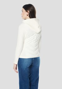 Fehér kapucnis pulóver steppelt, texturált hátsó panellel, kapucnival és hosszú ujjakkal. Kék farmernadrággal párosítva.