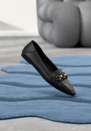 Mocassin en cuir noir avec détail en chaîne dorée posé sur un tapis bleu texturé à l'intérieur.