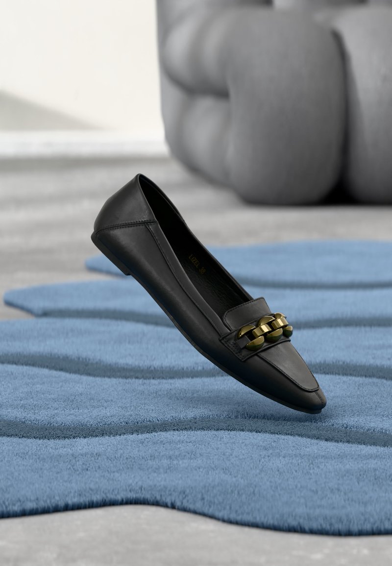 Mocassin en cuir noir avec détail en chaîne dorée posé sur un tapis bleu texturé à l'intérieur.