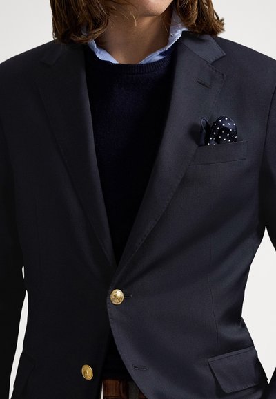 Blazer azul-marinho com botões dourados, combinado com um suéter escuro e uma camisa azul-claro. O lenço de bolso apresenta bolinhas brancas, acrescentando detalhe.
